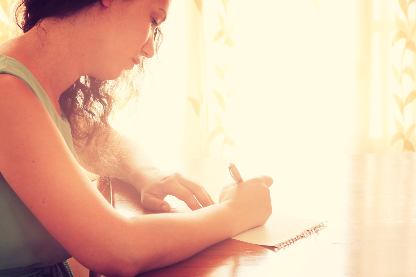 young woman sitting and writing letter near bright window light.