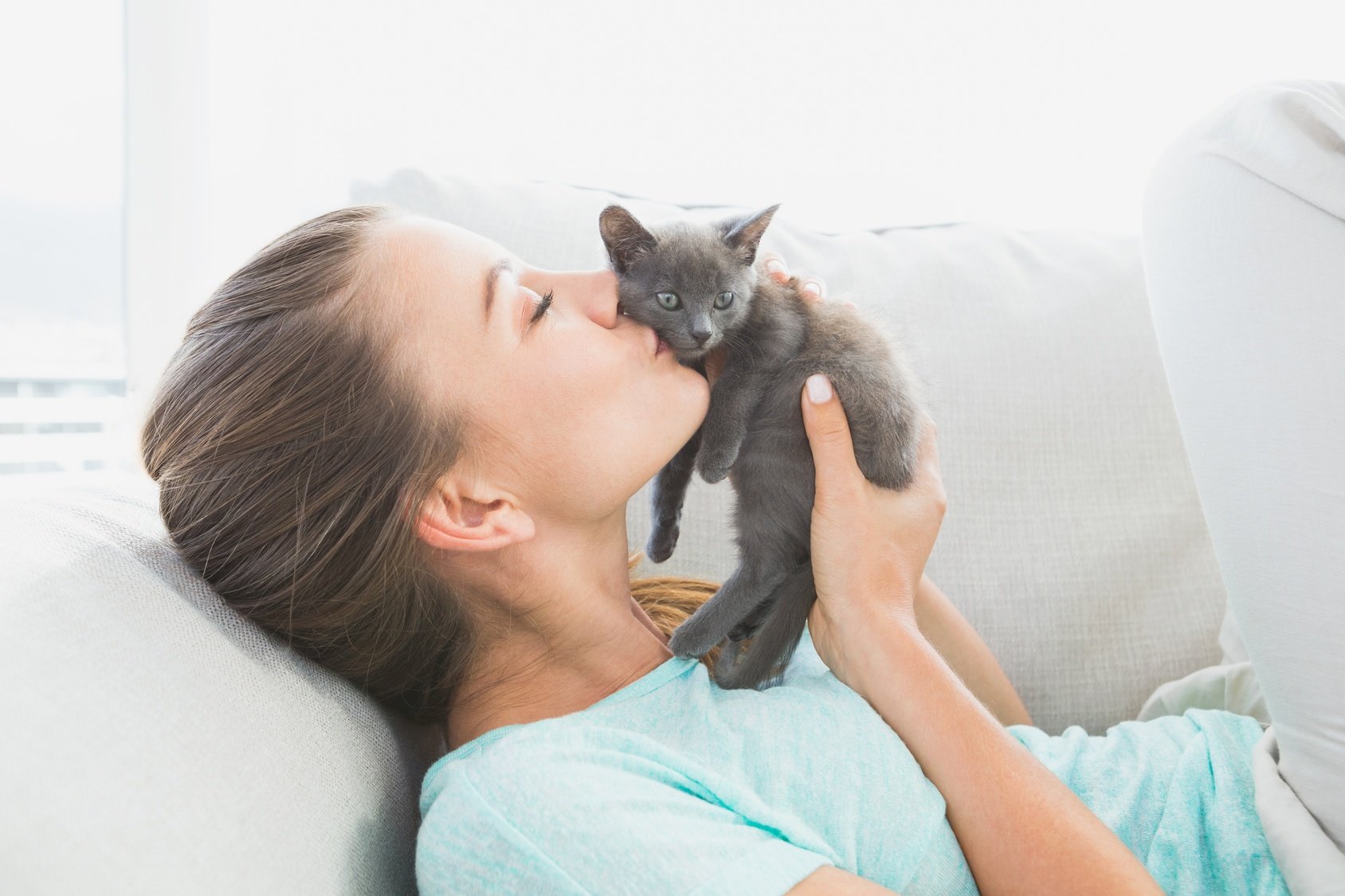 Cheerful woman lying on sofa kissing a grey kitten