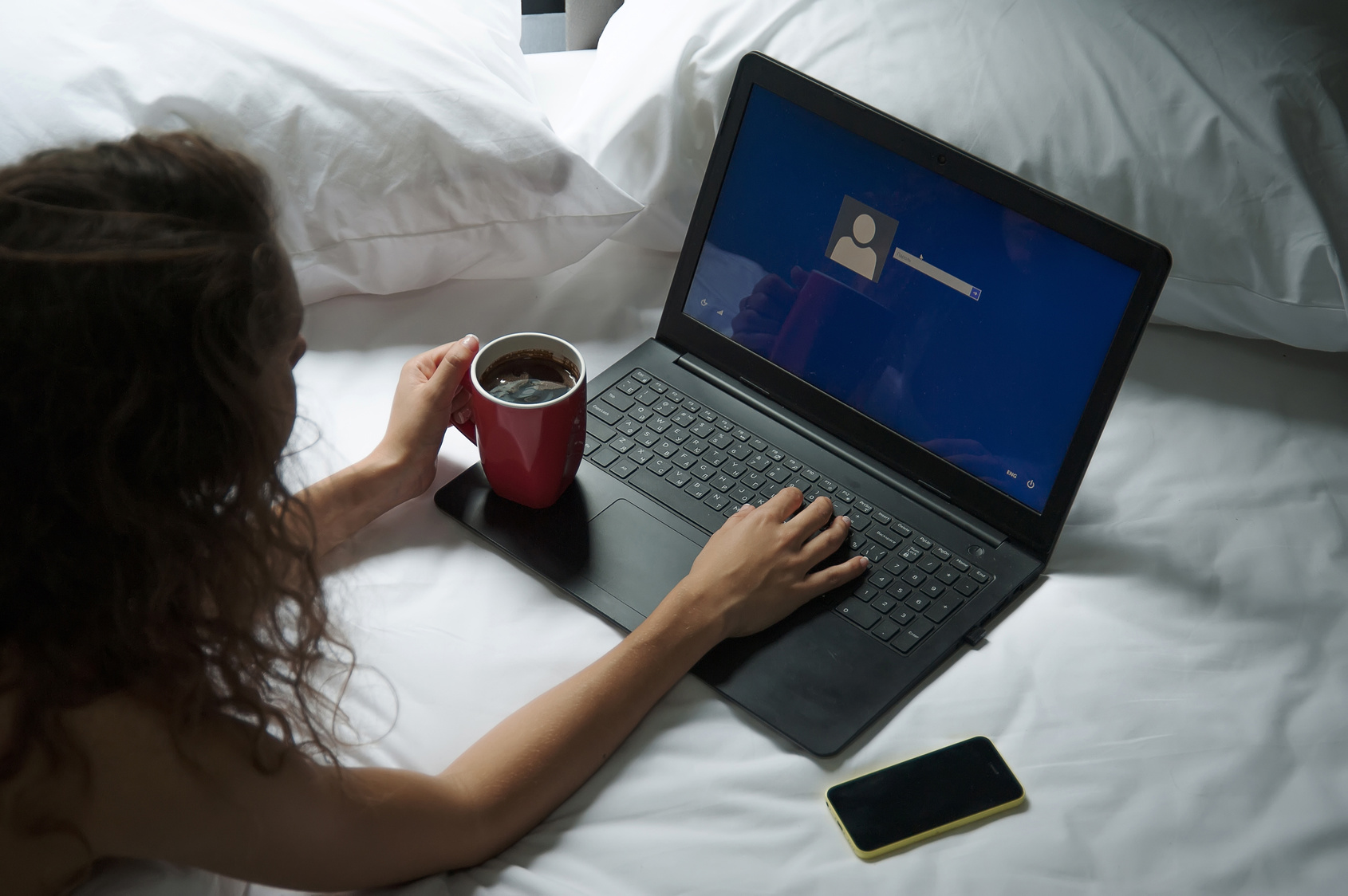 Woman is sitting on bed with a laptop and a cup of coffee