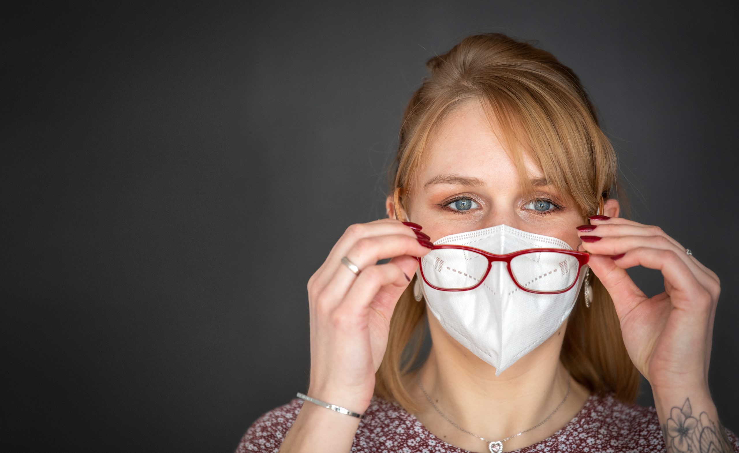 Problematisch: Mundnasenschutz und Brille – rotblonde Frau mit FFP2 Maske setzt ihre Brille auf – isoliert vor dunklem Hintergrund, Panoramaformat mit Textfreiraum