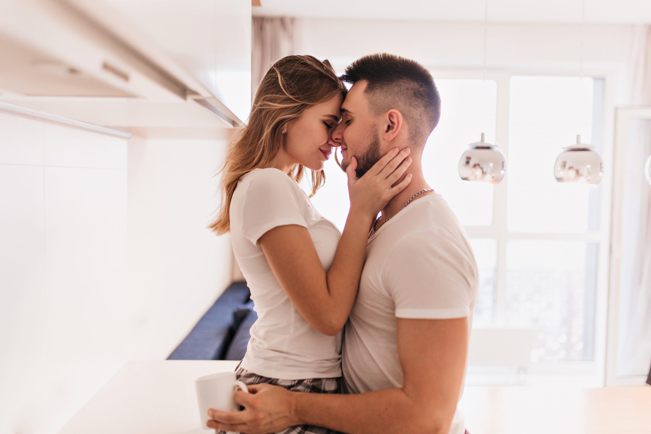 Sporty dark-haired man holding cup of tea and embracing wife. Enchanting girl in white attire gently touching boyfriend’s face..