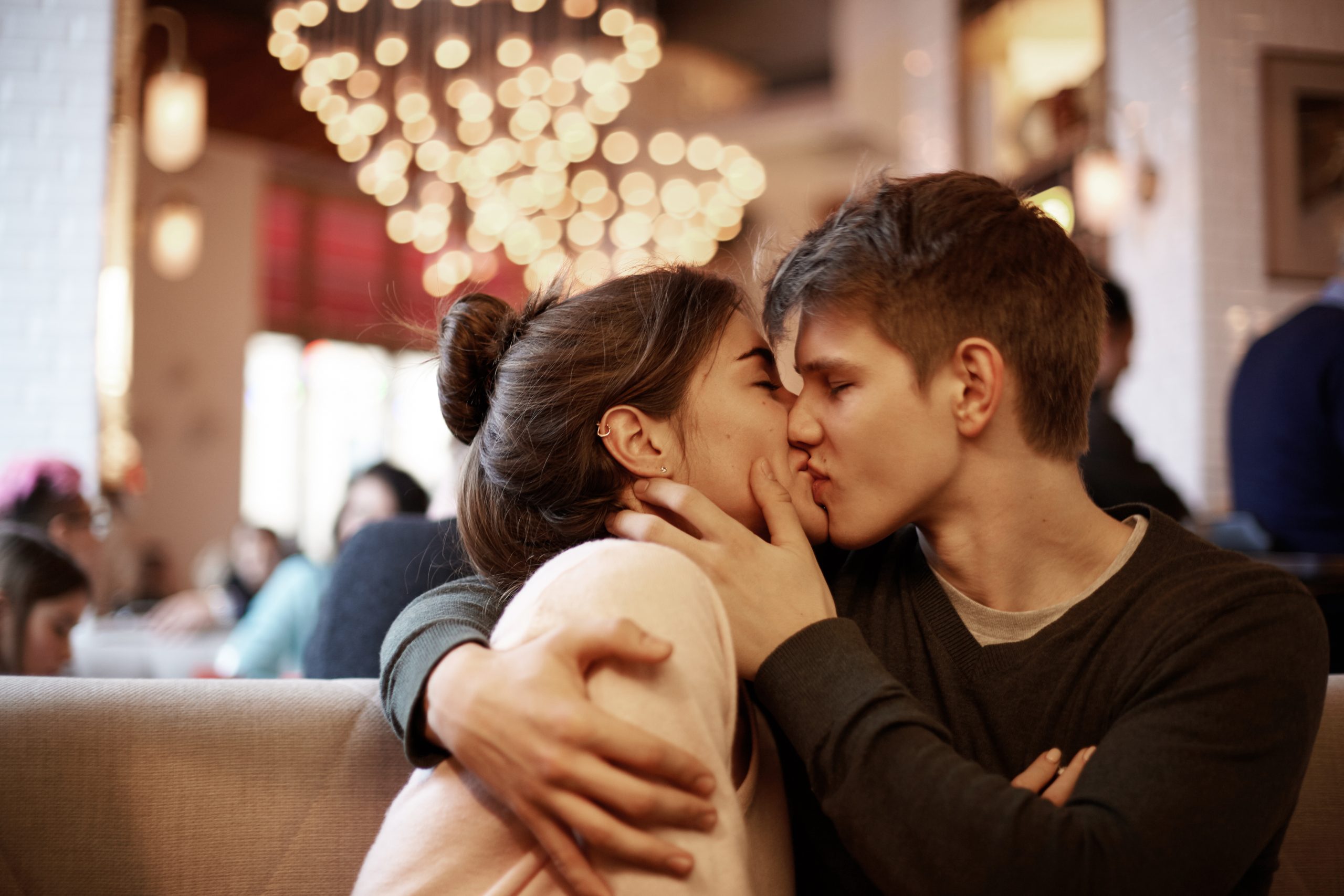 Candid shot of cute loving young couple relaxing at cafeteria on Valentine’s Day. Handsome guy holding tight his charming girlfriend and kissing her with fondness while waiting for lunch at cafe